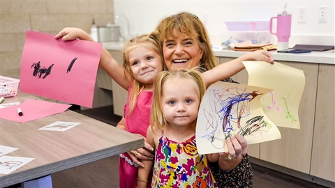 A smiling woman and two young girls are proudly holding their colorful artwork in a bright room with a kitchen background.