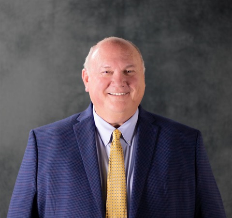A man in a blue checked suit and yellow patterned tie smiles warmly against a gray textured background. The mood is positive and professional.