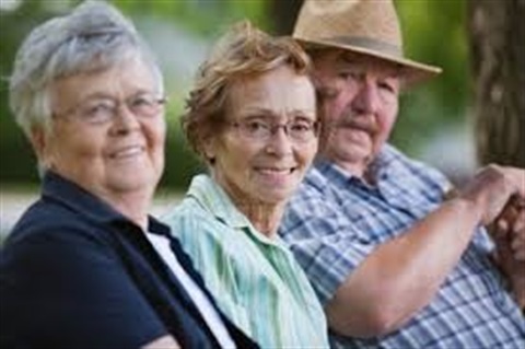 Three elderly people sit outdoors on a bench, smiling warmly. They appear relaxed, dressed in casual attire, with greenery blurred in the background.