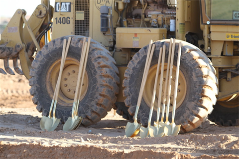 Seven golden shovels lean against the large tires of a yellow construction vehicle on a dirt ground, suggesting a groundbreaking ceremony.