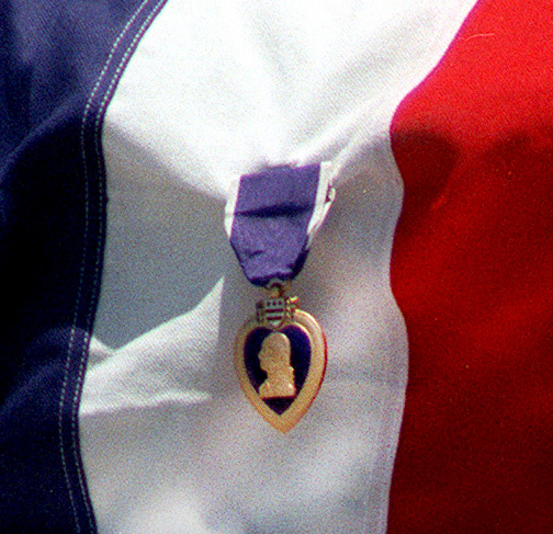 A Purple Heart medal hangs from a folded flag, featuring blue, white, and red colors. The medal is heart-shaped with a soldier's profile in the center.