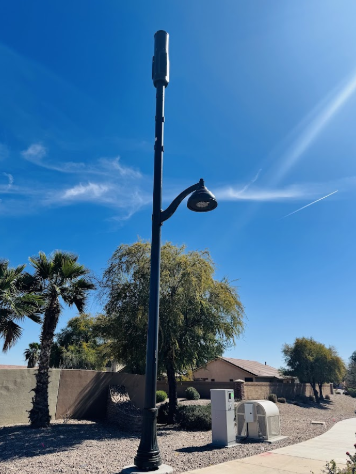 A black streetlight with a cellular antenna stands on a sidewalk near trees, houses, and utility boxes under a clear blue sky.