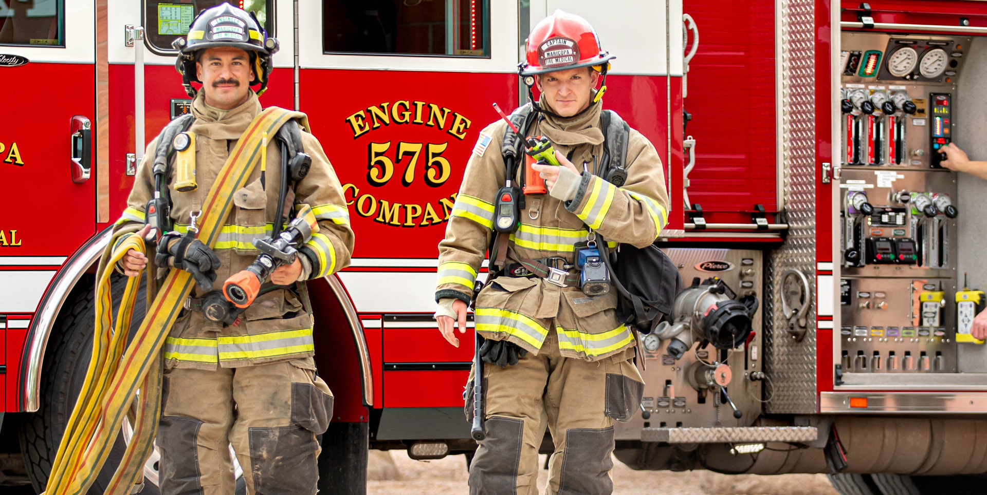 Two firefighters in full gear stand in front of a red fire engine labeled 