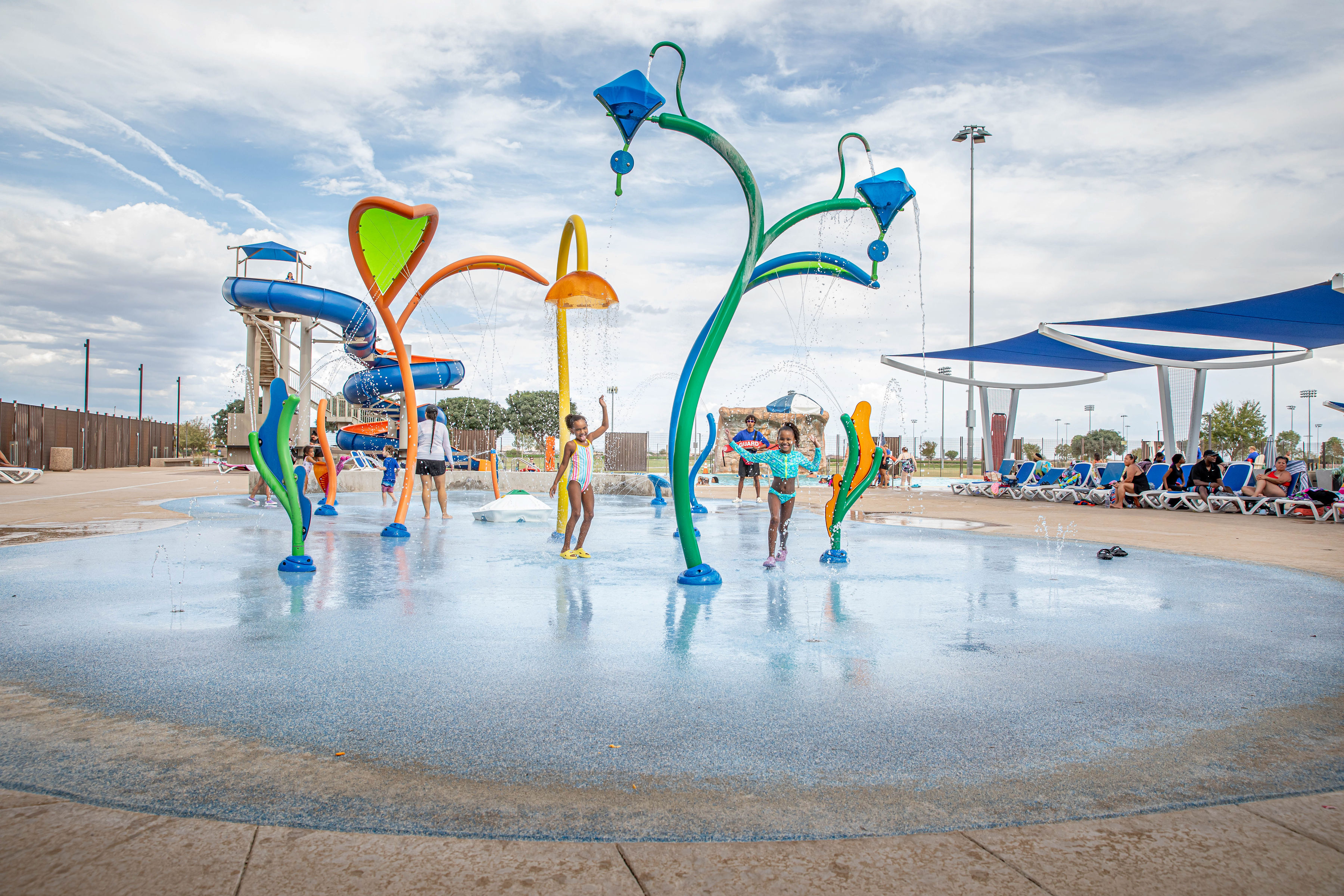Children playing joyfully at a colorful water park splash pad with vibrant fountains and slides on a sunny day, surrounded by people.