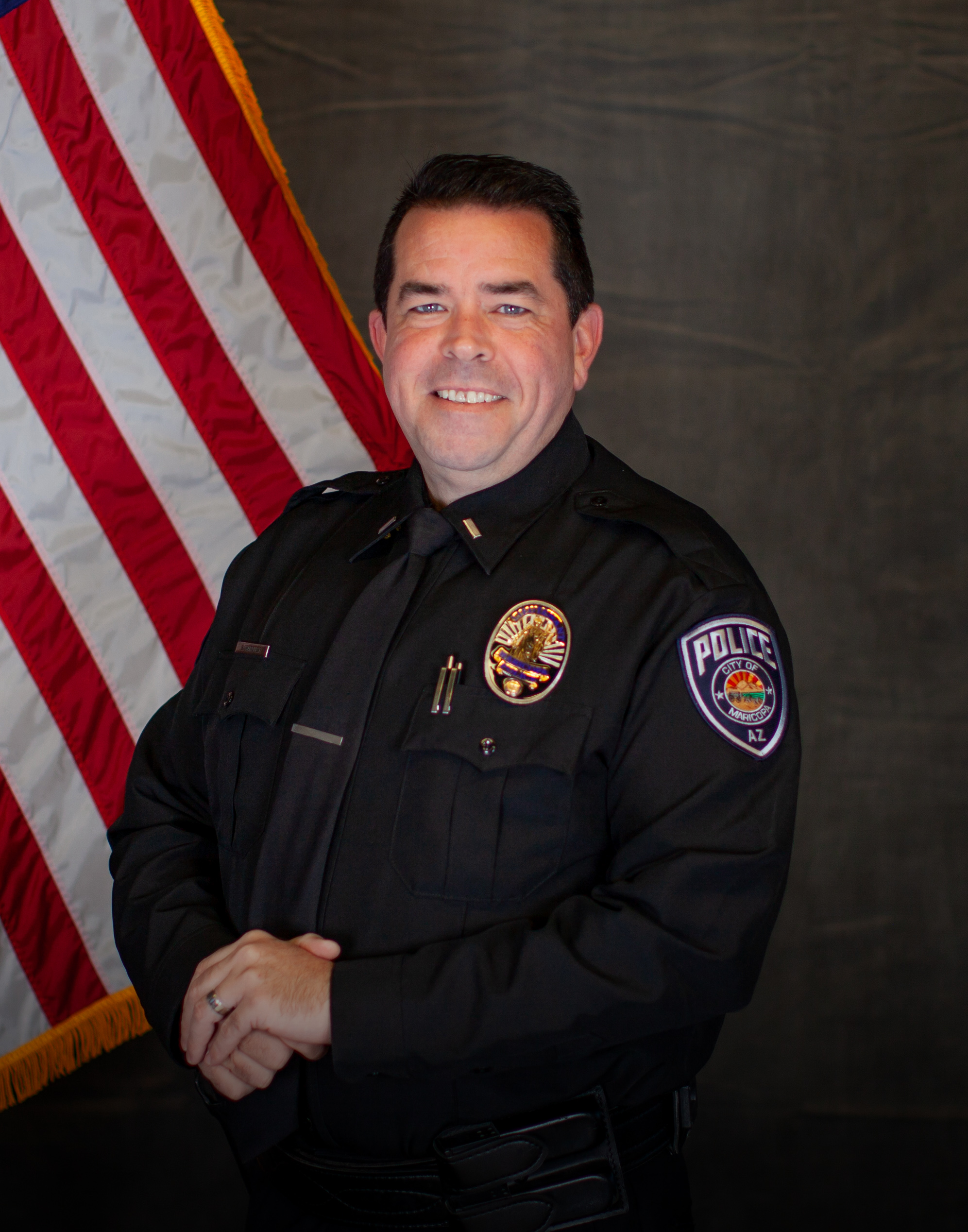 A smiling police officer in uniform stands proudly in front of a U.S. flag. The officer's badge and patch are visible, conveying authority and respect.