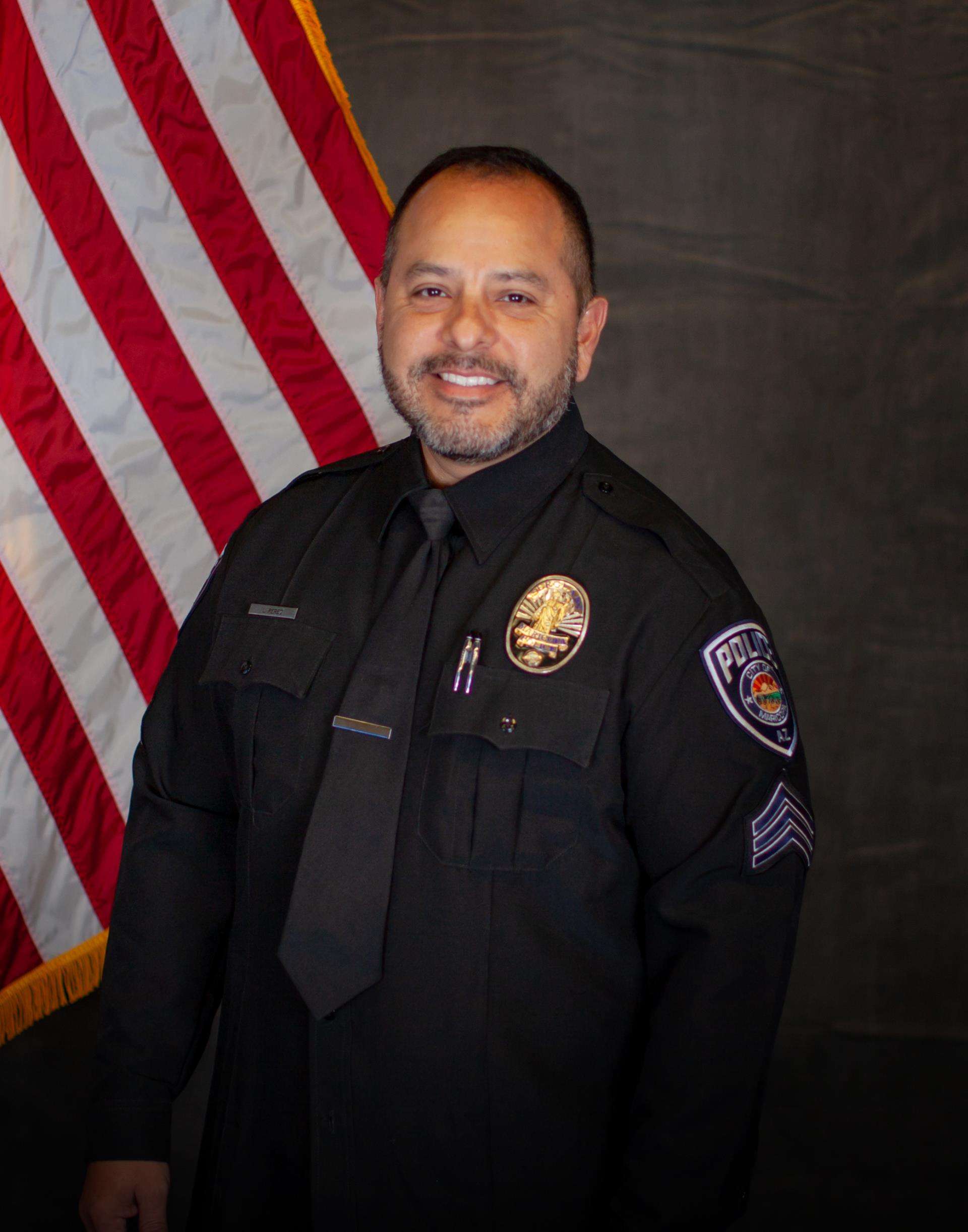 Police officer in a black uniform smiling confidently in front of an American flag. He wears a badge and has a calm, professional demeanor.