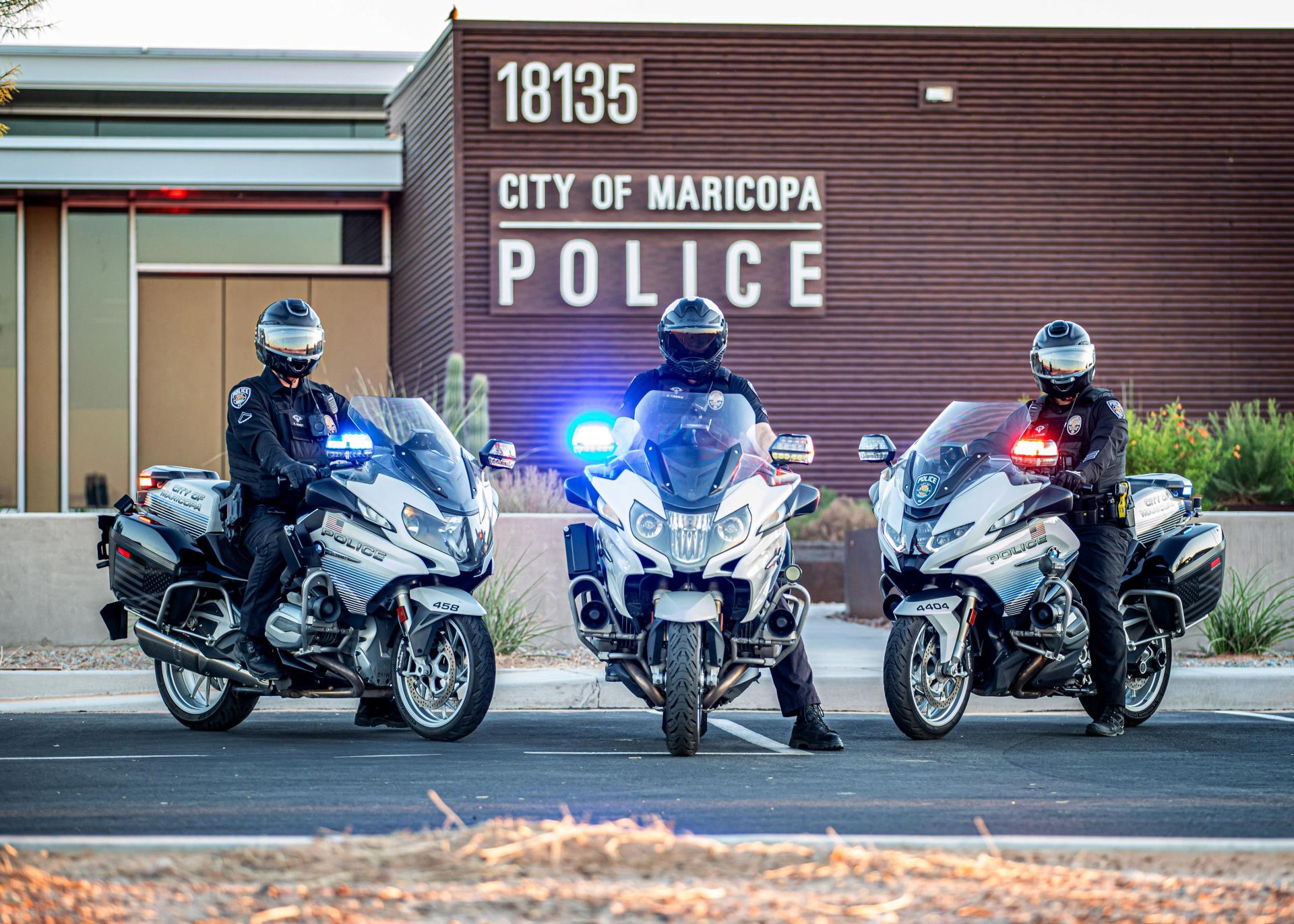 Three motorcycle police officers with helmets and uniforms are parked in front of the City of Maricopa Police Station. The motorcycles have flashing lights.