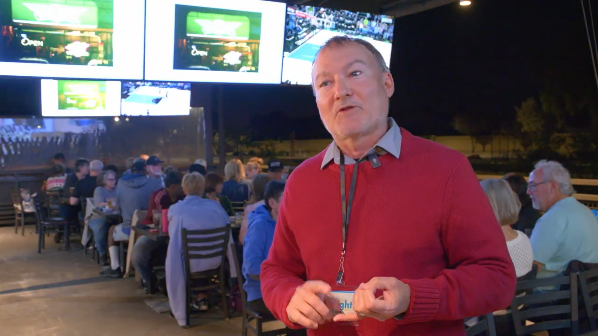 A man in a red sweater speaks in an outdoor venue, filled with people seated at tables. Multiple screens show a sports event, creating an energetic atmosphere.