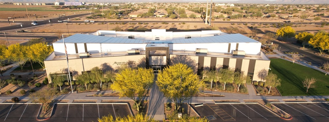 Aerial view of a modern building with a flat roof, surrounded by landscaped areas and parking lots. Trees with yellow leaves line the property. A mountain backdrop is visible.