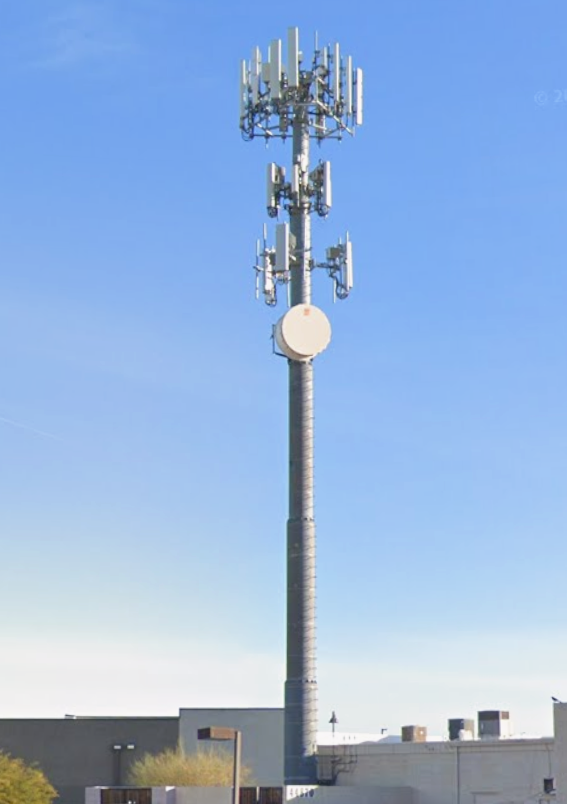 A tall cellular tower with multiple antennas stands against a clear blue sky, with part of a building visible at the bottom of the image.