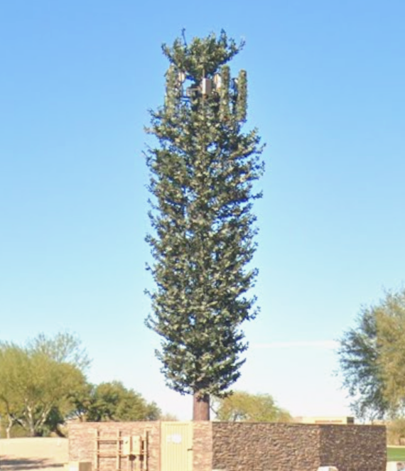 The image displays a tall, artificial tree structure designed to camouflage telecommunications equipment. The tree is slender and vertically elongated, resembling a pine tree with dense, green foliage achieved through realistic plastic branches. 