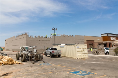 Construction scene with workers in hard hats atop an unfinished brick building. Vehicles, a container, and lumber are in the foreground. Clear sky above.
